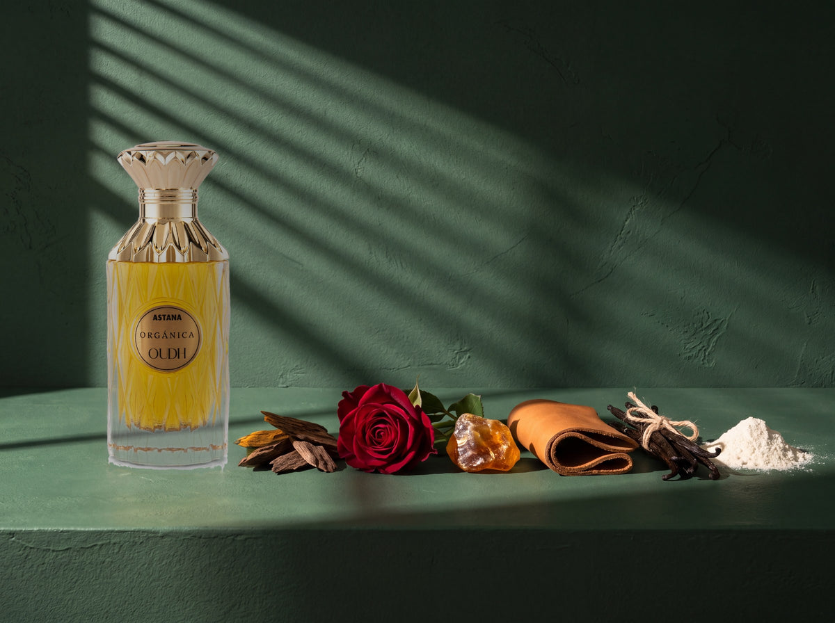 An Organica Oudh bottle standing on deep green plaster beside the six materials of the line: oud wood, a red rose, raw amber, tan leather, vanilla pods and white musk