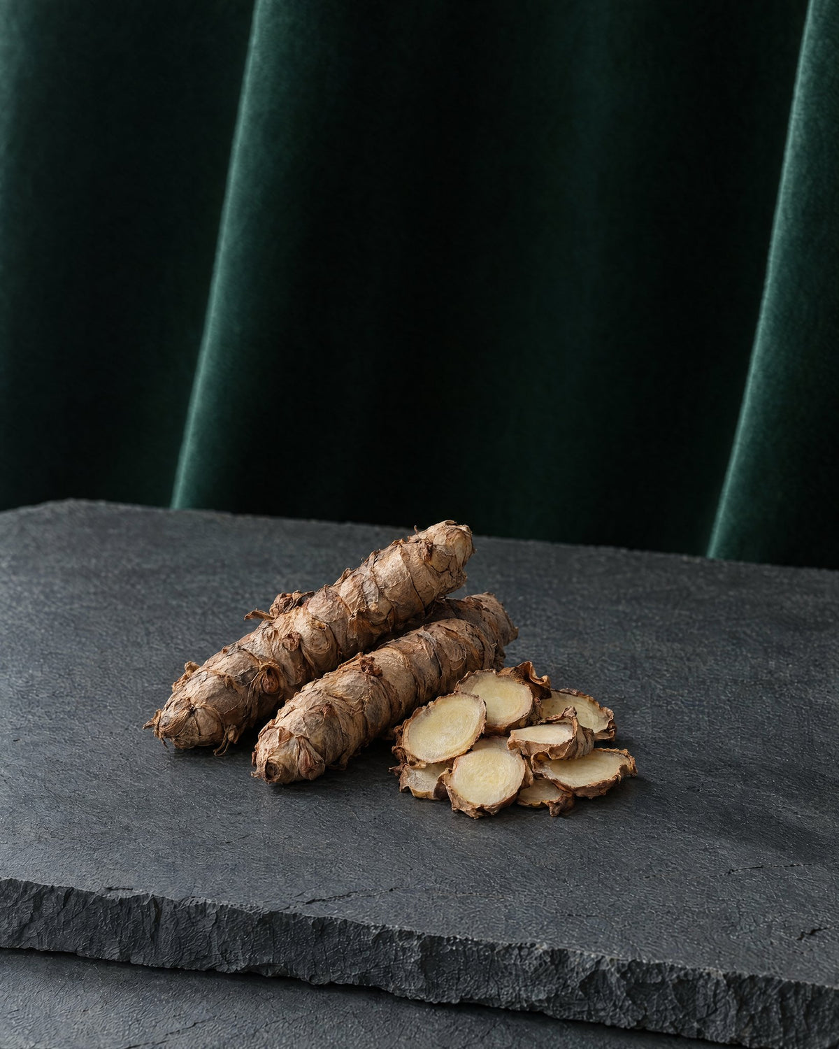 Kapur kachri rhizomes with papery brown skin, some sliced to show the pale flesh, on a slab of slate with deep green velvet behind