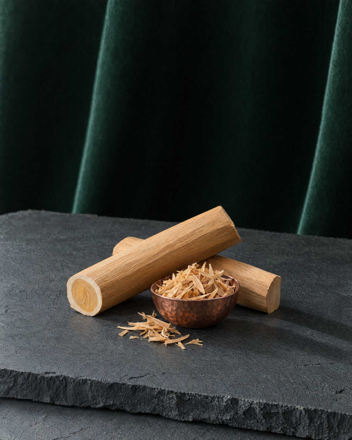 Two pale sandalwood billets and a copper bowl of fine shavings on a slab of slate, deep green velvet behind